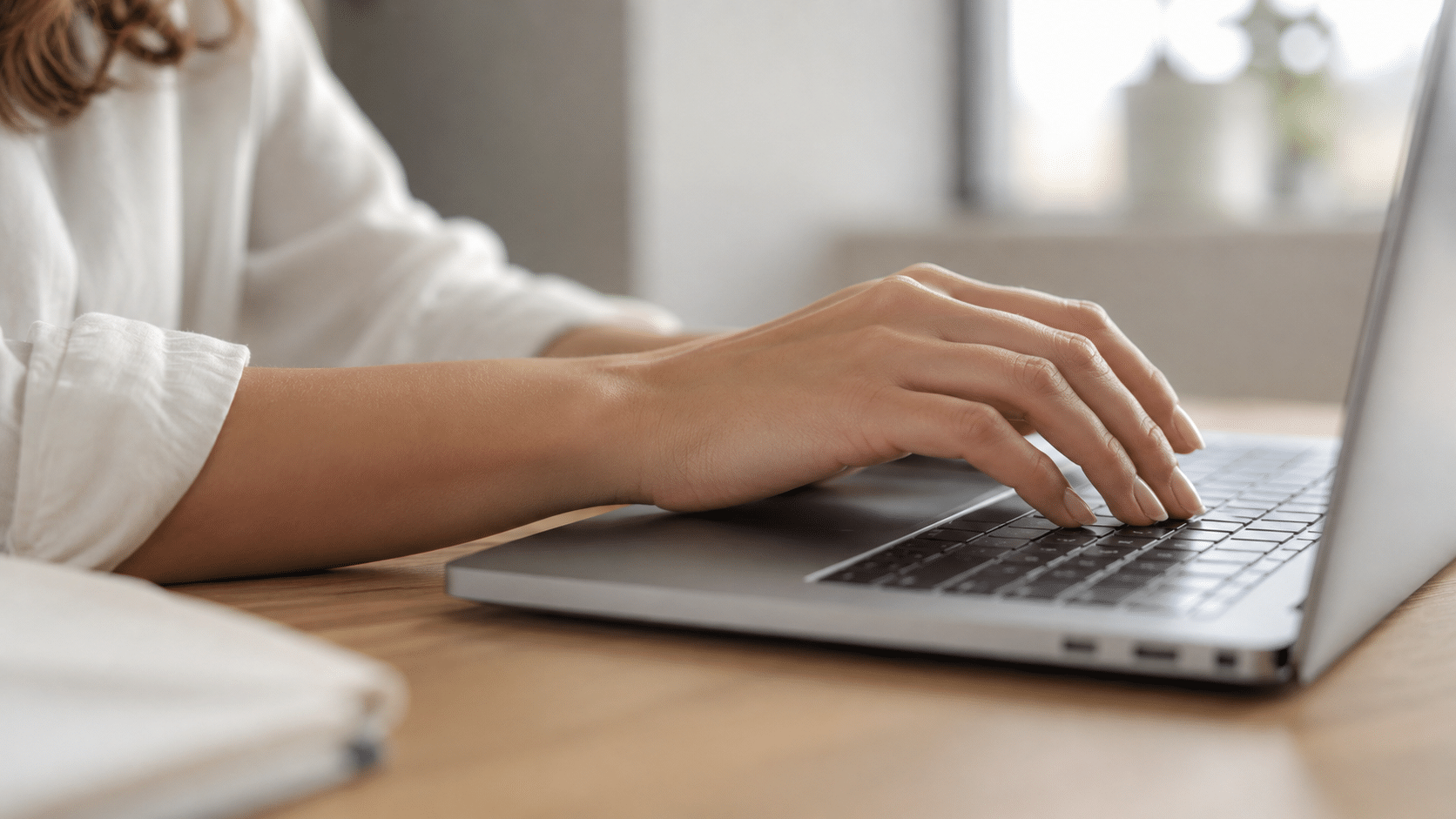 Mãos femininas digitando em laptop prateado sobre mesa de madeira clara.