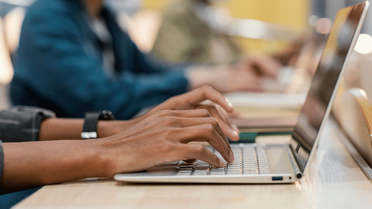 Duas pessoas digitando em notebooks lado a lado sobre mesa de madeira em sala de estudo.
