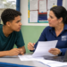 Estudante recebe orientação sobre inscrições do Sisu+ 2026 em ambiente escolar, com papéis e notebook sobre a mesa.