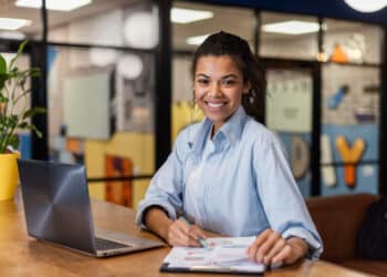 Jovem sorri com caneta e documentos ao lado do notebook durante a inscrição online
