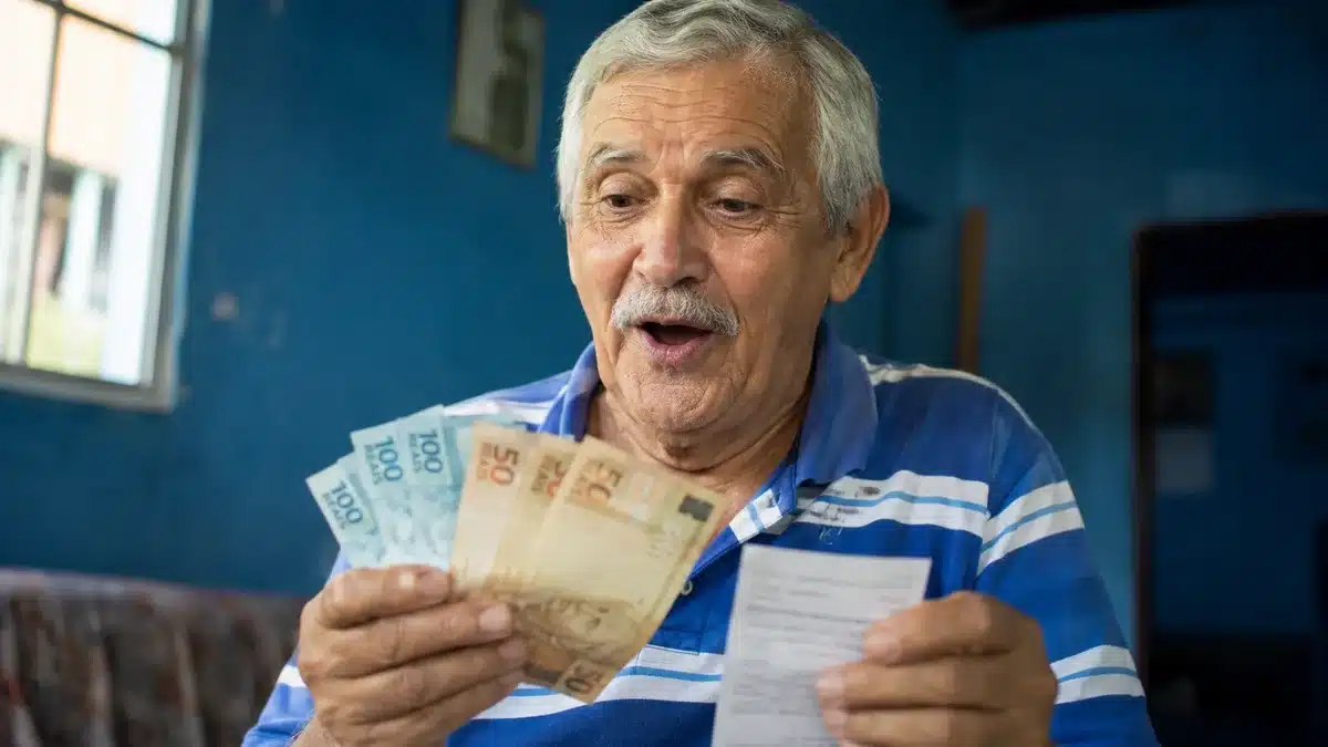 Aposentado brasileiro de cabelos grisalhos segurando cédulas de R$ 100 e R$ 50, olhando com expressão de surpresa, representando o pagamento de R$ 4.500 em 8 de junho.