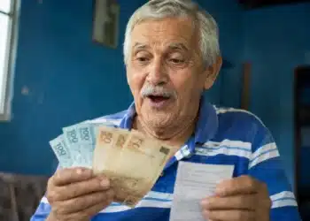 Aposentado brasileiro de cabelos grisalhos segurando cédulas de R$ 100 e R$ 50, olhando com expressão de surpresa, representando o pagamento de R$ 4.500 em 8 de junho.