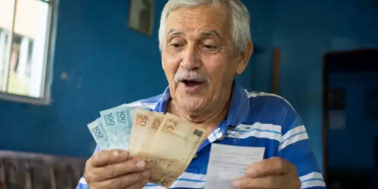 Aposentado brasileiro de cabelos grisalhos segurando cédulas de R$ 100 e R$ 50, olhando com expressão de surpresa, representando o pagamento de R$ 4.500 em 8 de junho.