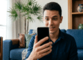 Homem sorri ao olhar para o smartphone enquanto está sentado na sala de casa