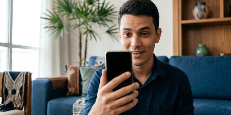 Homem sorri ao olhar para o smartphone enquanto está sentado na sala de casa