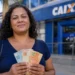 Mulher segura cédulas de real em frente a uma agência da CAIXA, representando a liberação do saque de R$ 6.220.