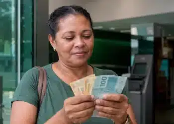 Mulher em agência bancária observa cédulas de reais com expressão de atenção, representando os pagamentos de R$ 946, R$ 1.351 e R$ 1.621 do PIS/Pasep.