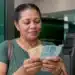 Mulher em agência bancária observa cédulas de reais com expressão de atenção, representando os pagamentos de R$ 946, R$ 1.351 e R$ 1.621 do PIS/Pasep.