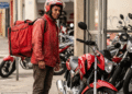 Entregador com mochila térmica observa moto vermelha, veículo que poderá ser financiado pela nova linha de crédito do governo