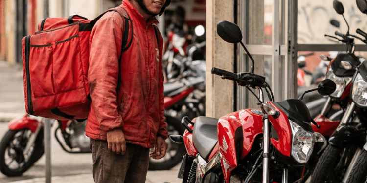 Entregador com mochila térmica observa moto vermelha, veículo que poderá ser financiado pela nova linha de crédito do governo