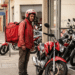 Entregador com mochila térmica observa moto vermelha, veículo que poderá ser financiado pela nova linha de crédito do governo