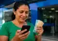 Mulher consulta o celular enquanto segura notas de real em frente a uma agência bancária, representando o pagamento do lote especial do Imposto de Renda via Pix.