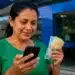 Mulher consulta o celular enquanto segura notas de real em frente a uma agência bancária, representando o pagamento do lote especial do Imposto de Renda via Pix.