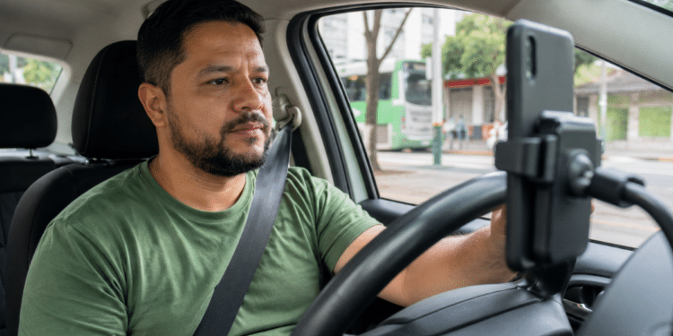 Motorista de aplicativo brasileiro dentro do carro utilizando smartphone no painel durante o trabalho.