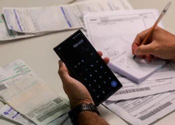 Pessoa calcula valores em celular enquanto organiza boletos e documentos de dívidas sobre a mesa.