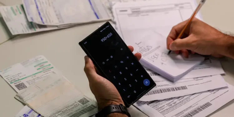 Pessoa calcula valores em celular enquanto organiza boletos e documentos de dívidas sobre a mesa.