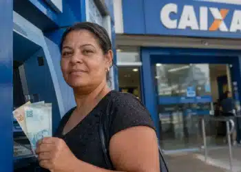 Mulher segura notas de reais em frente a caixa eletrônico da Caixa, representando o novo pagamento de R$ 6.220.