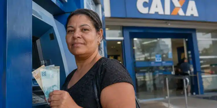 Mulher segura notas de reais em frente a caixa eletrônico da Caixa, representando o novo pagamento de R$ 6.220.