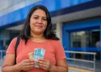 Mulher segura cédulas de real em frente a uma agência bancária azul, representando a nova liberação do saque-aniversário do FGTS.