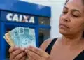 Mulher segura notas de real em frente a uma agência da Caixa, em referência ao pagamento do Bolsa Família de R$ 1.000 nesta quinta-feira (25).