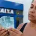 Mulher segura notas de real em frente a uma agência da Caixa, em referência ao pagamento do Bolsa Família de R$ 1.000 nesta quinta-feira (25).