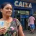 Mulher brasileira em frente a uma agência bancária da Caixa segurando dinheiro, representando o pagamento do Bolsa Família em junho.