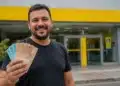 Homem sorridente segura cédulas de reais em frente a uma agência bancária amarela, representando o pagamento de até R$ 4.251 pela Caixa e pelo Banco do Brasil.
