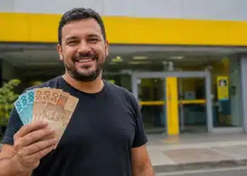 Homem sorridente segura cédulas de reais em frente a uma agência bancária amarela, representando o pagamento de até R$ 4.251 pela Caixa e pelo Banco do Brasil.