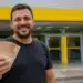 Homem sorridente segura cédulas de reais em frente a uma agência bancária amarela, representando o pagamento de até R$ 4.251 pela Caixa e pelo Banco do Brasil.