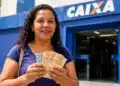 Mulher exibe cédulas de dinheiro em frente à agência da Caixa, representando a confirmação de pagamento de benefício no valor de R$ 6.220.