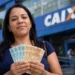 Mulher segura notas de real em frente a uma agência da Caixa, representando a confirmação do pagamento de R$ 800.