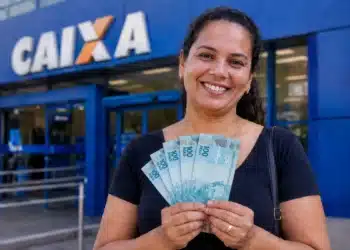 Mulher sorrindo segurando notas de R$ 100 em frente a uma agência da Caixa, representando o anúncio do pagamento de R$ 1.050.
