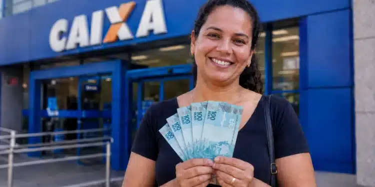 Mulher sorrindo segurando notas de R$ 100 em frente a uma agência da Caixa, representando o anúncio do pagamento de R$ 1.050.