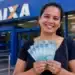 Mulher sorrindo segurando notas de R$ 100 em frente a uma agência da Caixa, representando o anúncio do pagamento de R$ 1.050.