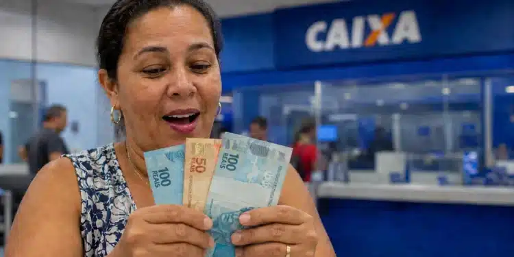 Mulher segura cédulas de real em agência da Caixa, representando o pagamento de R$ 2.900.