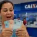 Mulher segura cédulas de real em agência da Caixa, representando o pagamento de R$ 2.900.
