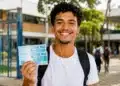Estudante com mochila sorri segurando cédulas de real em frente a uma escola, representando o pagamento do Pé-de-Meia em junho.