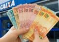 Mãos seguram cédulas diante de agência da Caixa, banco que paga os valores do Pé-de-Meia