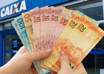 Mãos seguram cédulas diante de agência da Caixa, banco que paga os valores do Pé-de-Meia