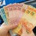 Mãos seguram cédulas diante de agência da Caixa, banco que paga os valores do Pé-de-Meia