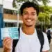 Estudante com mochila sorri segurando cédulas de real em frente a uma escola, representando o pagamento do Pé-de-Meia em junho.