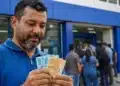 Trabalhador segura notas de R$ 50 e R$ 100 em frente a uma agência bancária, representando o pagamento de até R$ 3.746 do PIS/Pasep em junho.