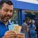 Trabalhador segura notas de R$ 50 e R$ 100 em frente a uma agência bancária, representando o pagamento de até R$ 3.746 do PIS/Pasep em junho.