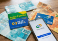 Cédulas de reais, cartões Bolsa Família e Pé-de-Meia com smartphone exibindo app Meu INSS sobre mesa de madeira.