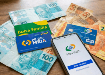 Cédulas de reais, cartões Bolsa Família e Pé-de-Meia com smartphone exibindo app Meu INSS sobre mesa de madeira.