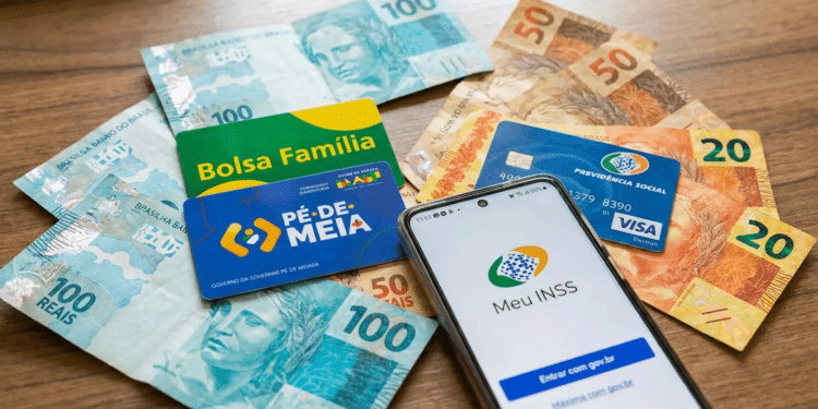 Cédulas de reais, cartões Bolsa Família e Pé-de-Meia com smartphone exibindo app Meu INSS sobre mesa de madeira.