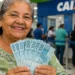 Idosa sorridente segurando notas de R$ 100 em agência bancária, representando o pagamento de R$ 8.475,55 do INSS em junho.