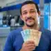 Homem brasileiro sorrindo, segurando cédulas de R$ 100 e R$ 50, representando os pagamentos de R$ 406, R$ 675 e R$ 946 que serão liberados em 15 de junho.