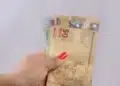 Mão segurando notas de real em destaque, representando pagamentos do programa Pé-de-Meia no novo ciclo de benefícios.