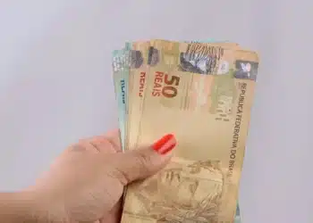 Mão segurando notas de real em destaque, representando pagamentos do programa Pé-de-Meia no novo ciclo de benefícios.